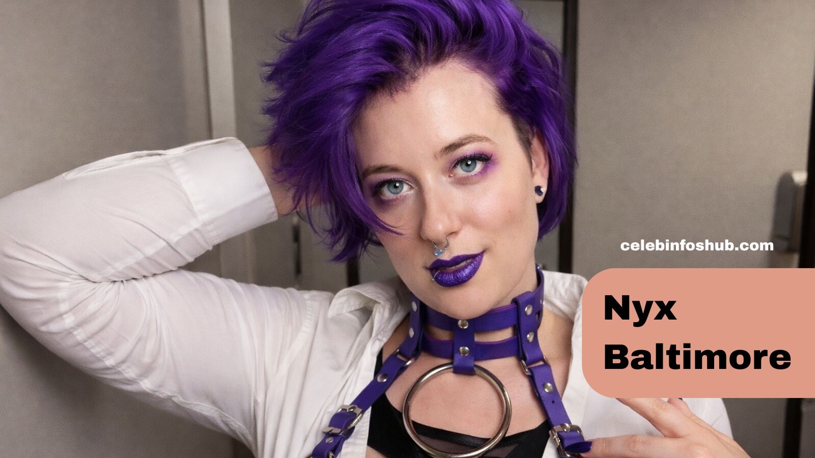 Portrait of a confident woman with short vibrant purple hair, bold purple lipstick, and statement piercings, posing indoors against a neutral background. Stylish and edgy fashion look featuring a white shirt and purple harness accessories, inspired by the bold aesthetic of Nyx Baltimore.